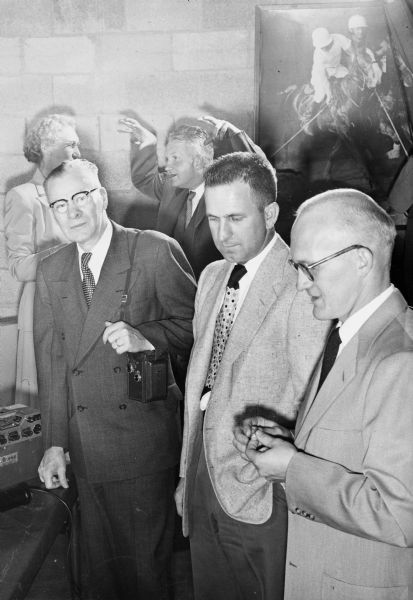 Slightly elevated view of three well-dressed men standing together in the foreground, with the man on the left holding a camera over his shoulder. Behind the three men, Sid is standing with a cigar in his mouth talking to a woman and making gestures with his hands up in the air. On the wall behind Sid is a photograph or print of two men on horseback playing polo. On the left on a table is an unidentified machine.
