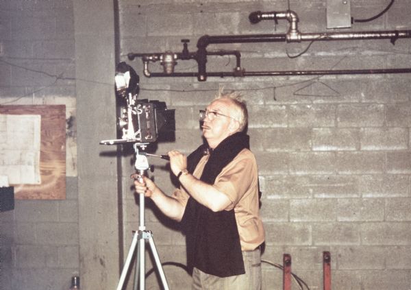 Sid is standing and operating a camera on a tripod inside a building with concrete walls and exposed pipes. He is wearing eyeglasses and has a cigar in his mouth. A black focusing cloth is around his neck. 