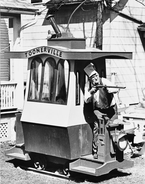 Portrait by photo journalist, Edwin Stein, of Sid in his backyard standing on the back of the Toonerville Trolley holding a shotgun. Sid is wearing a large engineer-style hat and has a cigar or unknown object clamped in his mouth. The back porch of the house in on the left.