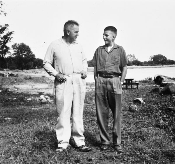 Outdoor portrait of Sid and his son, Steve Boyum, standing on the grass with a picnic table and what may be a lake in the background.
