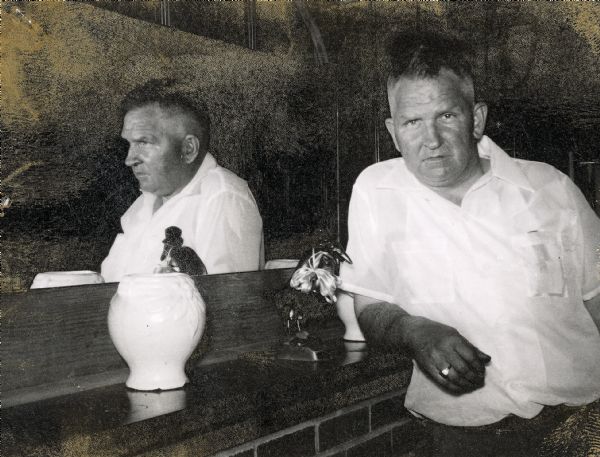 Dual portrait of Sid, <i>en face</i> staring straight at the camera and in profile reflected in a large mirror. He is leaning with his arm on a long shelf that frames the mirror. He wears a white shirt and a ring on his ring finger. The mantel is decorated with a vase and a small bird sculpture.