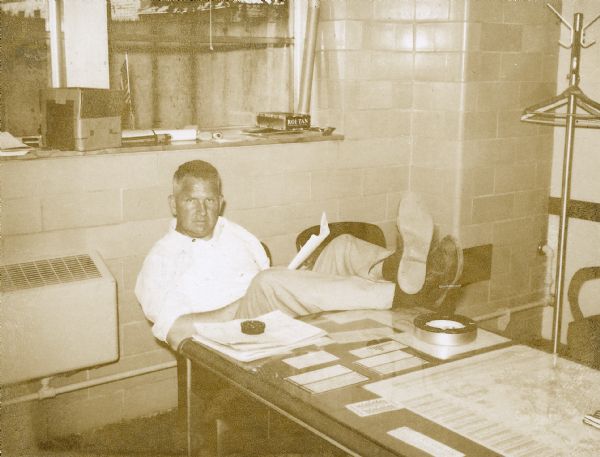 Portrait of Sid sitting in a chair holding work papers with his legs up on a large desk in front of him, probably at Gisholt. The desktop has maps and notes pressed under a large sheet of glass, and a stack of papers and large round ashtray on top. The office has walls covered in large plain glazed tiles, two windows, and a standing coat rack. 