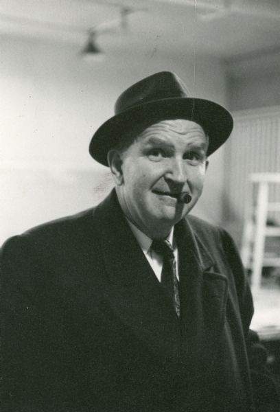 Waist-up portrait of Sid wearing an overcoat and hat, with his signature cigar in his mouth.