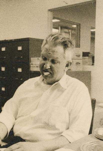 Waist-up portrait of Sid sitting in an office at a desk or table. He has a cigar in his mouth. Behind him are standing file cabinet and boxes of supplies. 