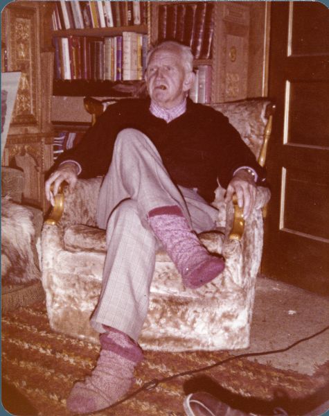 Portrait of Sid sitting in an armchair at his home at 237 Wabesa Street, wearing a pair of socks. He has a cigar in his mouth. Behind Sid is a bookshelf.