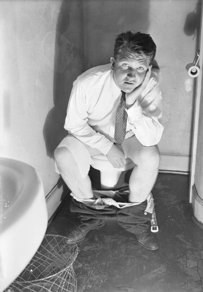 Self-portrait of Sid Boyum using a slave unit for the flash. He is wearing a shirt and necktie sitting on a toilet with his pants around his ankles. He is resting his head in his left hand. There is a piece of toilet paper over his right knee. 