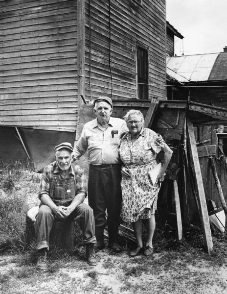 Outdoor group portrait of Sid, with a cigar in his mouth, posing with his arms around a man, sitting on the left, and a woman standing on the right. They are standing near the side of a building. Possibly Sid's Boyum grandparents.