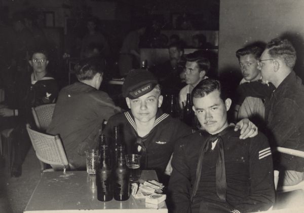 Portrait of two young U.S. Navy sailors sitting at a table in a tavern. There is a pack of Lucky Strike cigarettes and a book of matches in front of the mustached man, and a single, smoking cigarette on the edge of the table by the other. Cold bottles of beer stand in front of them. Other men are sitting at tables in the background. 