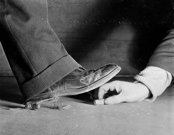 Close-up of a man's shoe treading on someone's hand holding a cigar butt on the ground, with a shadow casting on the wall in the background. On the ground near the cuff of the pants are two pine cones and pine needles. The "smoke" from the cigar appears to be retouched.