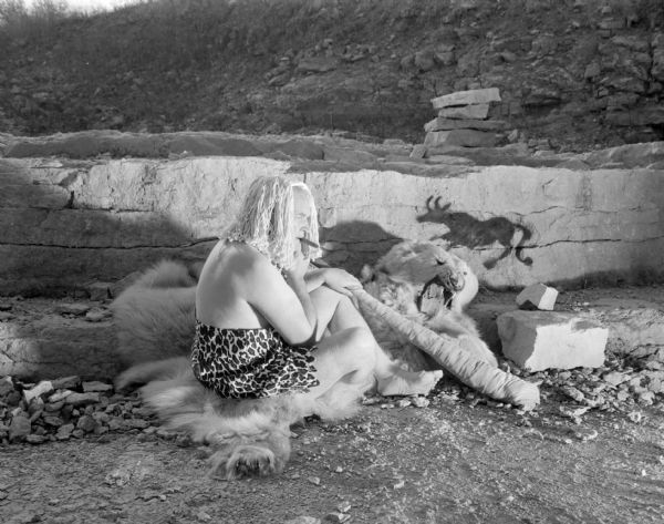 View of Sid, with a cigar in his mouth and holding a club, sitting on an animal-skin rug outdoors in what may be a quarry. He is wearing a rag mop on his head, and a faux animal skin. A faux cave painting of an animal is on one of the quarry walls in the background.