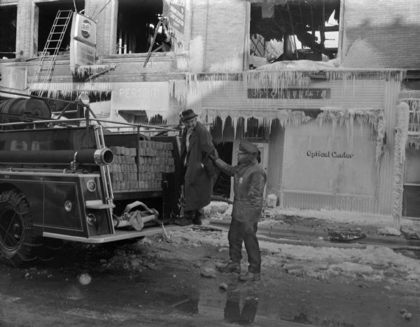 View across street towards Sid standing on the back of a fire engine with a cigar in his mouth. A police officer wearing a uniform and a badge is standing in the street behind Sid and is supporting Sid's arm. In the background are storefronts destroyed by fire and covered with icicles. There are signs for Pepsi-Cola and a Delicious Shish Kebob restaurant, and an "Optical Center."