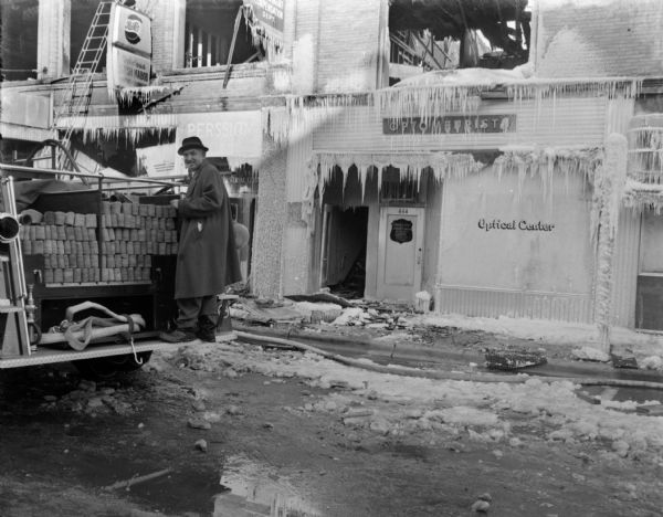View across street towards Sid standing on the back of a fire engine with a cigar in his mouth. In the background are storefronts destroyed by fire and covered with icicles. There are signs for Pepsi-Cola and a Delicious Shish Kebob restaurant, and an "Optical Center."