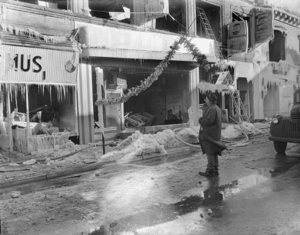 View of Sid standing in the street with a cigar in his mouth looking at damaged storefronts covered with icicles. Snow and ice cover the sidewalk, and a fire hose is snaking along the sidewalk. The storefront windows are broken, and a truck, perhaps a fire engine, is parked on the far right. There are signs for Pepsi-Cola, a Delicious Shish Kebob restaurant, and the Industrial Commission.