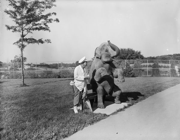 View across sidewalk towards Sid standing and holding a shovel while posing next to an elephant sitting on a bench outdoors. Sid is wearing a white coat, white hat and boots. There is a chain-link fence in the background.