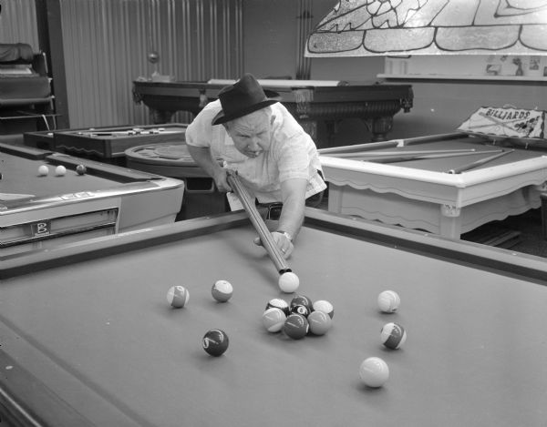 Sid bending over a pool table in a game room using a shotgun as a pool cue. He is wearing a hat and has a cigar in his mouth.