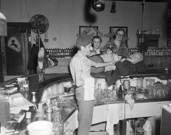 View across curved bar towards Sid and three other men at the Karabis tavern. Sid is laying on top of the bar on his back, with his head resting on a blanket and a cigar in his mouth. Sid is holding a glass in his left hand, and two men standing behind him are resting glasses of beer on his stomach. The bartender is standing inside the bar on the left, holding Sid's left wrist, perhaps taking his pulse.
