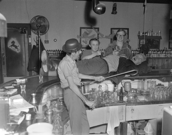 View across curved bar towards Sid and three other men at the Karabis tavern. Sid is laying on top of the bar on his back with his eyes closed, his hands folded on his chest, his head resting on a blanket and a cigar in his mouth. Two men standing behind Sid are resting glasses of beer on his stomach. The bartender is standing inside the bar on the left wearing a plastic hat and holding a cigarette in one hand and a cane in the other.