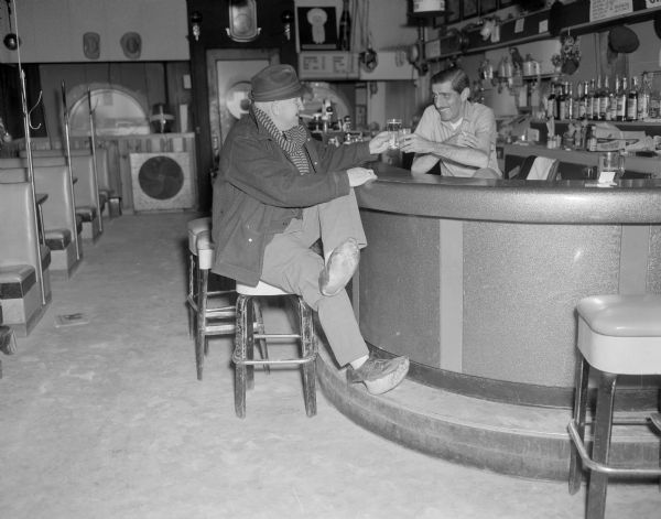 Sid, wearing a hat, coat, and scarf, and with a cigar in his mouth, is sitting on a bar stool at the Karabis tavern, toasting with the owner standing behind the bar. The bottoms of Sid's shoes are gone. 