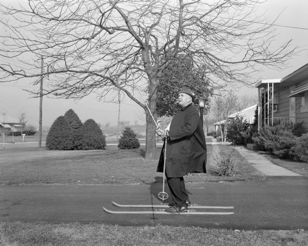 View from lawn towards Sid standing on skis in a driveway, lifting up one of the ski poles. Sid is wearing a coat, hat, and shoes, and has a cigar in his mouth. Houses are in the background.