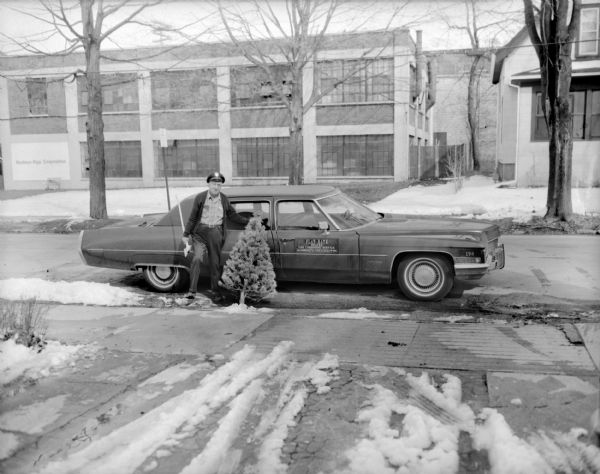 View down driveway towards Bob Bender posing standing at the curb leaning against a Cadillac Fleetwood limousine holding a small Christmas tree and an axe. Across Waubesa Street is a Madison-Kipp Corporation factory building. Snow is on the ground.