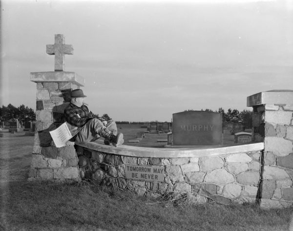 View of Sid posing sitting with his feet up on the stone wall at the entrance to a cemetery with a July 1961 calendar under his arm and a cigar in his mouth. He is looking off to the right towards the gravestones in the background. A sign on the wall just below Sid reads: "Tomorrow May Be Never." 