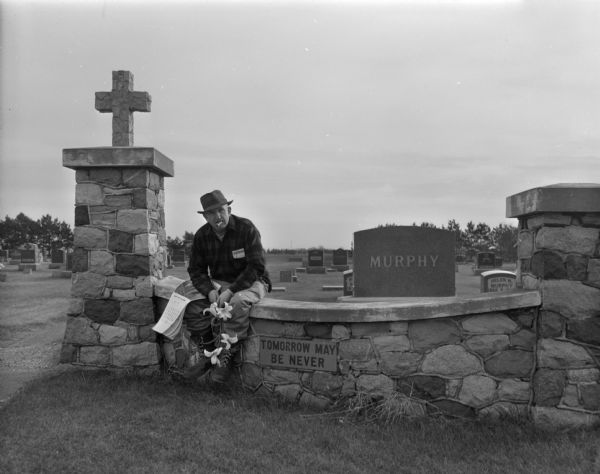 View of Sid posing sitting on the stone wall at the entrance to a cemetery with a July 1961 calendar under his arm. He is holding a stem of lilies and has a cigar in his mouth. There are gravestones in the background. A sign on the wall just below Sid reads: "Tomorrow May Be Never." 