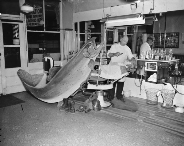 A man (Sid?) sitting in a barber's chair is getting a straight-edge shave while holding a fishing rod with a large cutout of a giant fish caught on the fishing line. On the floor at the base of the chair are a pair of boots, a creel, a fishing hat, and a box of fishing lures. The sign outside of the large front window of the entrance reads in part: ". . . mation . . . about trolling . . . booked here . . . for charter."