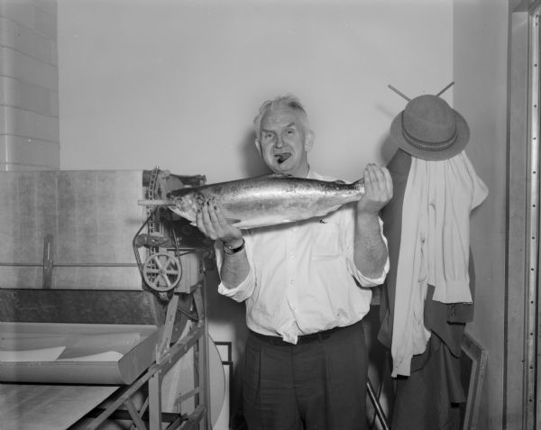 Portrait of Sid standing and holding up a trout in his hands. Both Sid and the trout have an unlit cigar in their mouths. Sid's nails are stained, probably with pyrogallol, a staining agent used in the development of film. A photographic print washer is on the left, and a coat and hat are on a coat rack on the right.