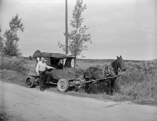 View across road towards Sid standing next to a horse-drawn vehicle, which appears to have been made from an old automobile. In the background is a field.