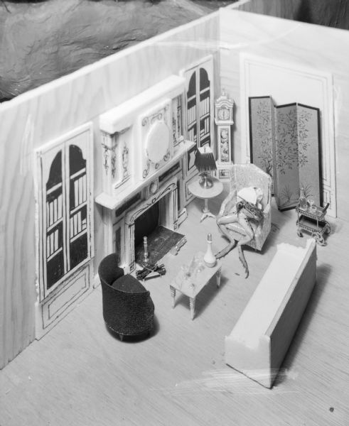 View looking down at a diorama of a living room with doll-sized furniture. A frog is sitting in a wing-back chair next to a fireplace.