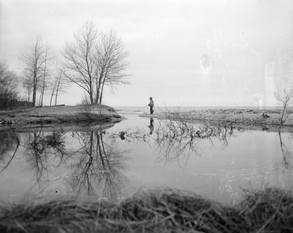 View from shoreline across water towards a man standing and fishing at the narrow inlet of a large body of water in the background. The man is wearing a hat and is smoking a pipe. Trees along the bank on the left are bare of leaves.