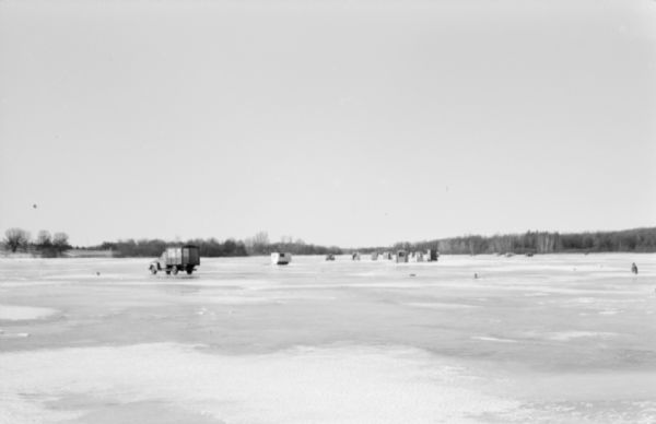 View across frozen lake towards cars parked on the ice near fishing huts. A shoreline lined with trees is in the far background.