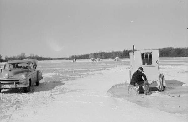 Sid Boyum ice fishing on a lake. He is sitting on a chair near an ice fishing hut. On the left a car is parked with the trunk open. Other ice fishing huts and cars are in the far distance.