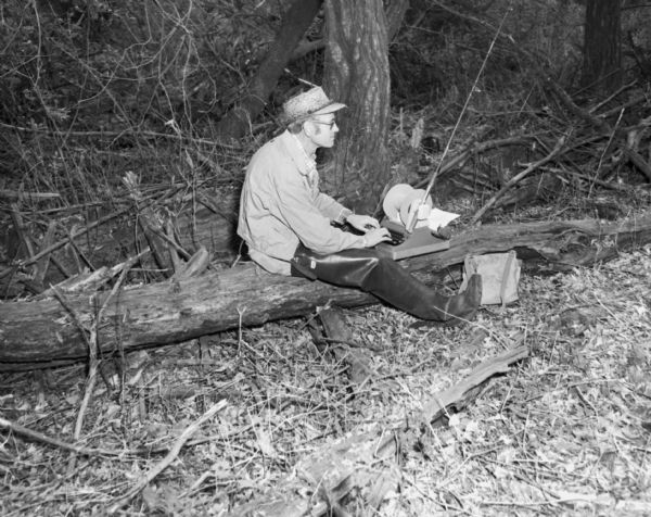 A man wearing a straw hat, coat, eyeglasses, and hip waders is sitting on a log typing on a typewriter. A fishing pole is propped up on top of the typewriter.