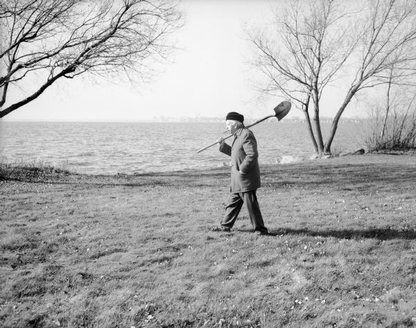 Sid, wearing an overcoat and a hat, is strolling along the Lake Monona shoreline with a cigar in his mouth. He is carrying a shovel over his right shoulder and has his left hand in his pocket. The Madison skyline is in the far background.