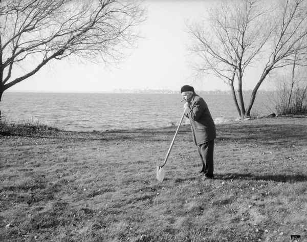 Sid, wearing an overcoat and a hat, is standing along the Lake Monona shoreline with a cigar in his mouth. He is leaning with both hands on a shovel propped on the ground. The Madison skyline is in the far background.