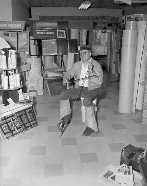A man sitting on plastic buckets in a store selling floor materials pretending to play a fishing pole with a bow. The man is wearing hip waders pulled down over his lower legs.