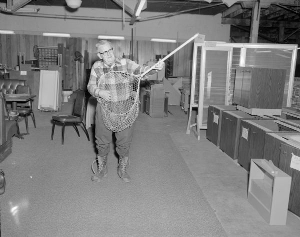 A man is standing and pretending to play a fishing net like a guitar. He is inside a large room crowded with furniture and stacked cabinetry.