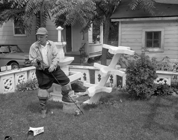 Sid standing with a fishing pole in his backyard. He is wearing folded down hip waders and he has a cigar in his mouth. He is standing with his foot propped up on an upside down bucket in the grass. Behind him are two of his sculptures. A fence surrounds the yard he is standing in, and in the background a car is parked near a house. In the foreground on the grass are a pair of shoes, and a photograph of Sid standing with a horse-drawn vehicle made out of an old automobile.