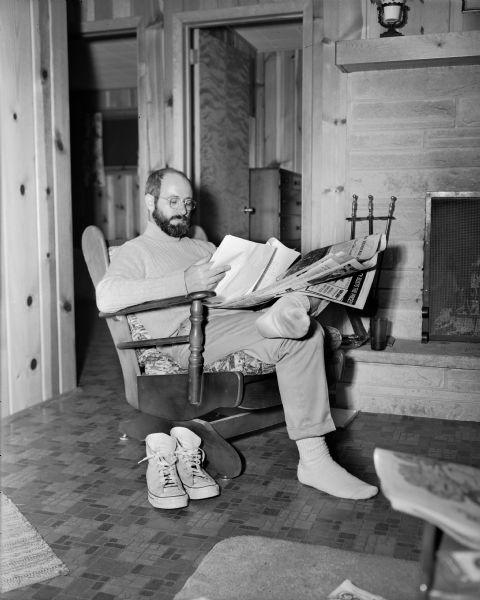 Steve Hopkins, the outdoor writer for the <i>Wisconsin State Journal</i>, sits in an armchair reading a manuscript next to a fireplace in his cabin. There is also a newspaper in his lap.