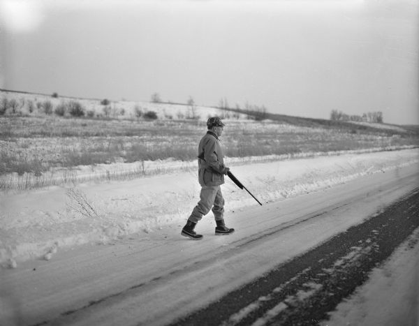 Al Dvorak with a shotgun walking on a road. Snow is on the ground.