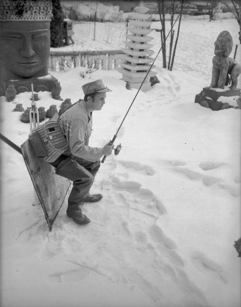 A man posing with a fishing pole in his hand is bending over while leaning against a board in Sid's snowy backyard. He is wearing hip waders, a hat, and a creel. Sculptures by Sid are in the background. In the background automobiles are parked in a lot.