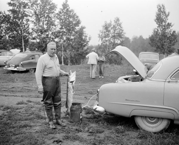 A man wearing hip waders is standing and holding a Northern Pike in his hand near the open trunk of a Packard Ultramatic. People are standing in the background near other parked cars.
