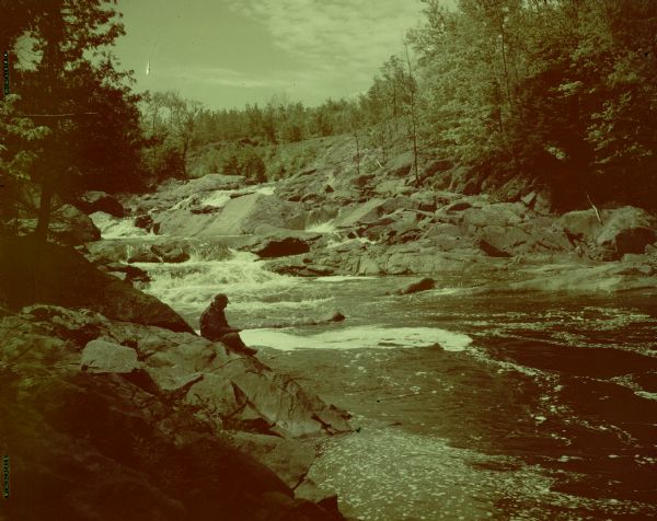 View along rocky shoreline towards Sid. He is sitting on a rocky outcropping and fishing in Bad River. Behind him are waterfalls.