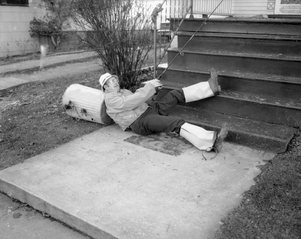 View of Stan Hamre,  laying along the concrete sidewalk at the foot of Sid Boyum's front porch steps, with one of his legs resting on one of the steps. He is partially leaning on an overturned garbage can and is holding up a fishing rod. He is wearing a hat, eyeglasses, and hip waders folded over his boots.