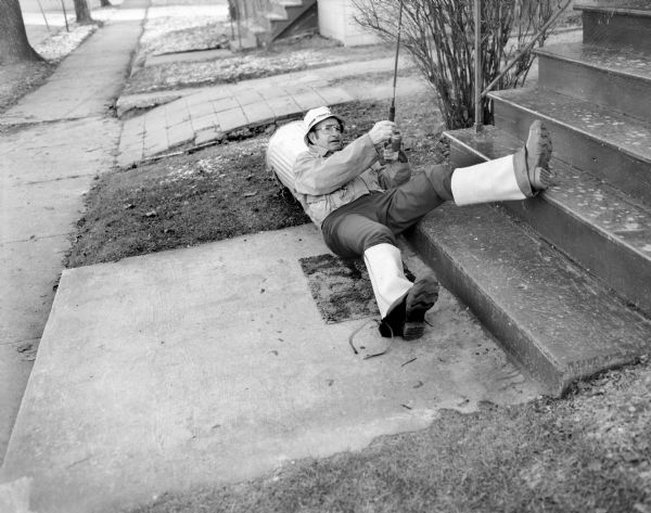 View of Stan Hamre laying along the concrete sidewalk at the foot of Sid Boyum's front porch steps, with one leg resting up on the steps. He is partially leaning against an overturned garbage can and is holding up a fishing rod. He is wearing a hat, eyeglasses, and hip waders folded over his boots.