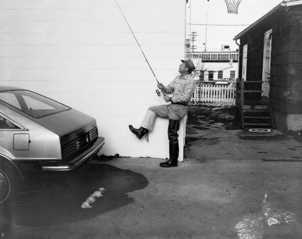 View of Carl Vogt leaning against the corner of a concrete block building holding up a fishing rod and raising his right leg. He is wearing hip waders, with one of the waders folded down over his boot. A Pontiac Phoenix is parked on the left, and in the far background down an alley behind a fence are cars parked in a lot in front of a factory building.