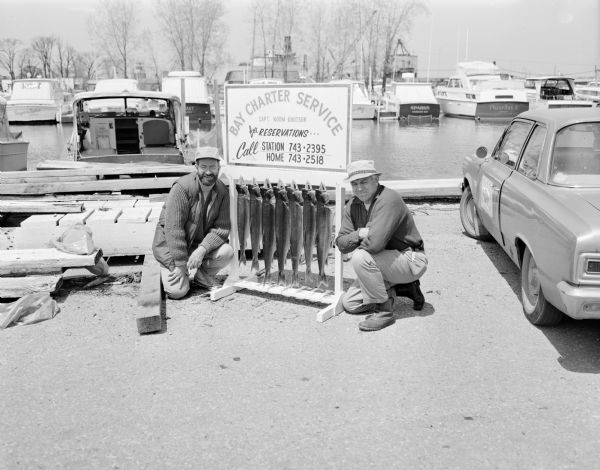 Steve Hopkins and Roger Debenham posing in Sturgeon Bay with seven trout they caught on Lake Michigan hanging from pegs under a sign that reads, "Bay Charter Service Capt. Norm Knutson." Behind them are boats moored in a harbor.