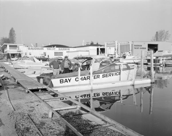Steve Hopkins and two men standing on a Bay Charter Service boat docked at the Baudhuin Yacht Harbor marina in Sturgeon Bay. The yacht club house stands behind them, and they are surrounded by other boats in slips.