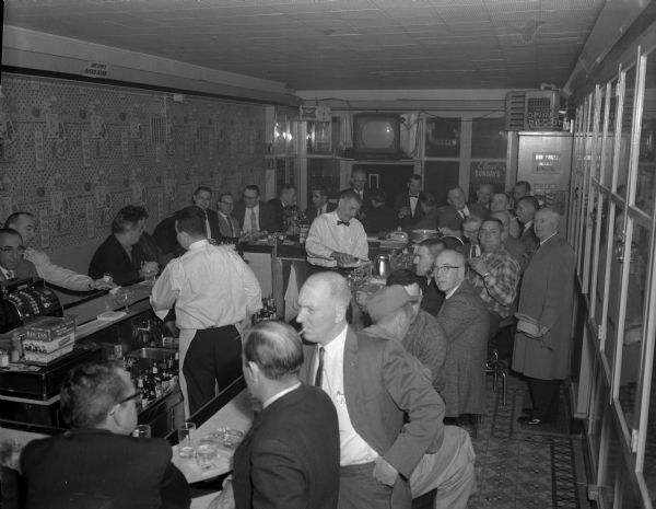 Elevated view of a crowded bar, all men, in the evening before a fishing banquet. Two men are bartending behind the rectangular shaped bar. A sign high up on the wall opposite the bar reads, in upside down letters: "This Sign Is Upside Down."
